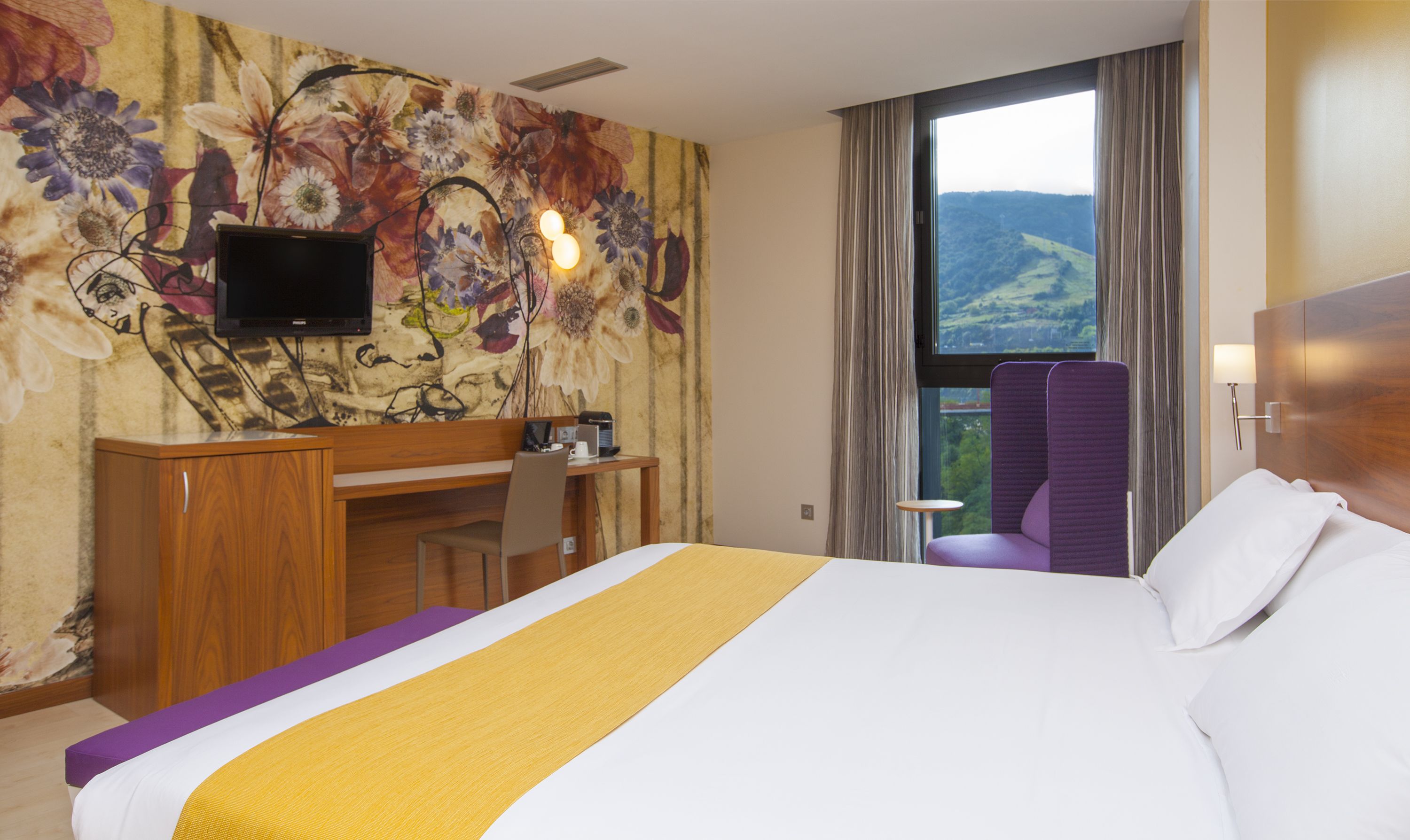 Une chambre d’hôtel équipée d’un lit, d’un bureau, d’une chaise, d’une télévision et d’une fenêtre donnant sur la campagne.