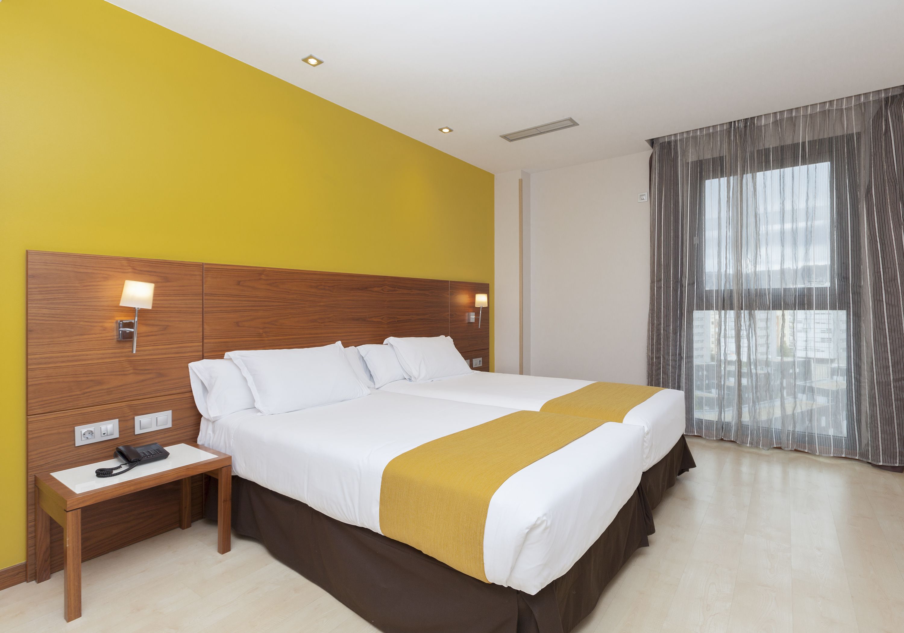 Ein Hotelzimmer mit einem großen Bett, einem Holzkopfteil, Nachttischlampen, einem Telefon und einem Fenster mit durchsichtigen Vorhängen.