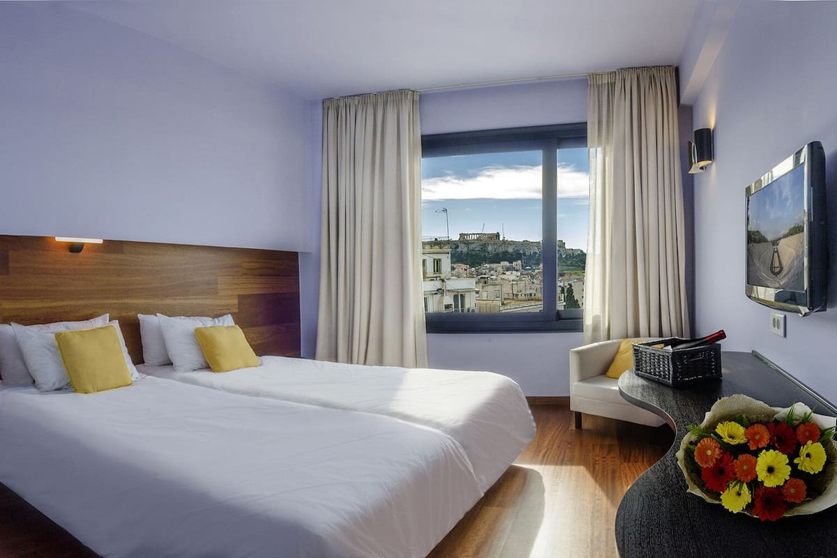 A hotel room with two beds, a view of the city, a desk with a chair, and a vase of flowers.