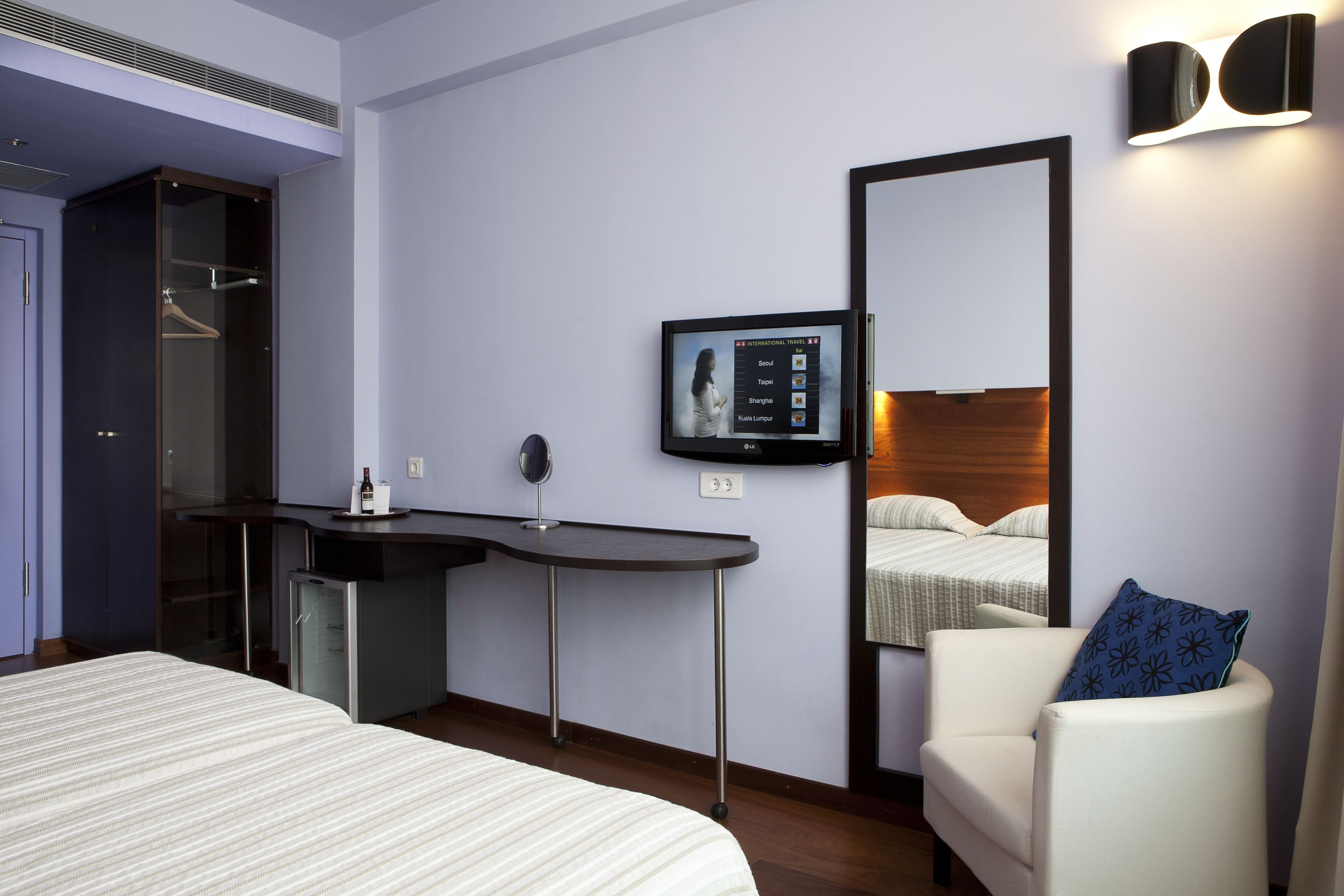 Une chambre d’hôtel équipée d’un lit, d’un bureau, d’une chaise, d’un miroir et d’un téléviseur affichant une application de voyage.