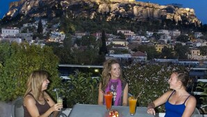 Three people seated at an outdoor table with drinks, overlooking a cityscape with a prominent hill and buildings. - Athens Center Square Hotel (Athens)