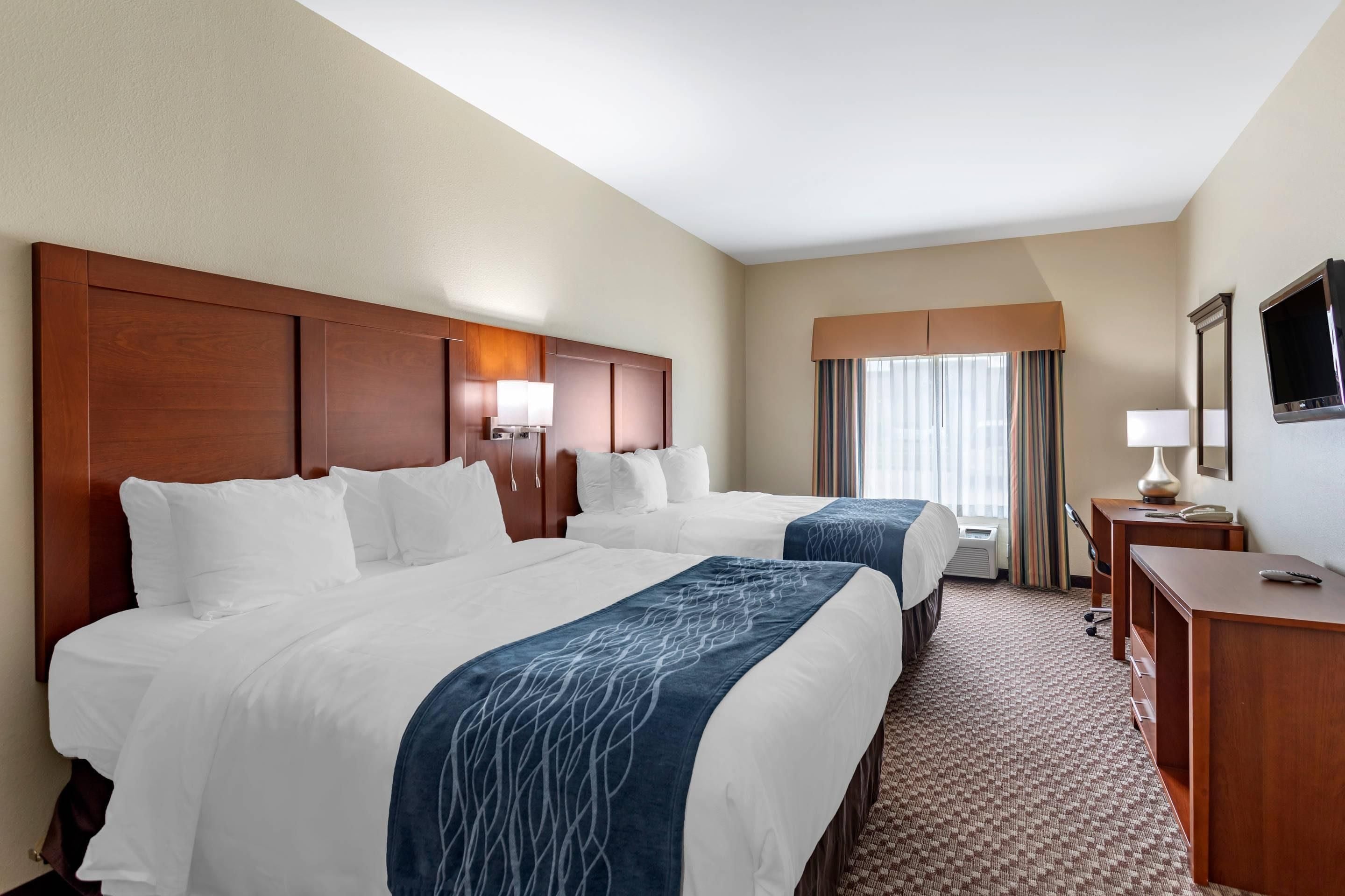 A hotel room with two beds, a desk, a TV, and a window with curtains.