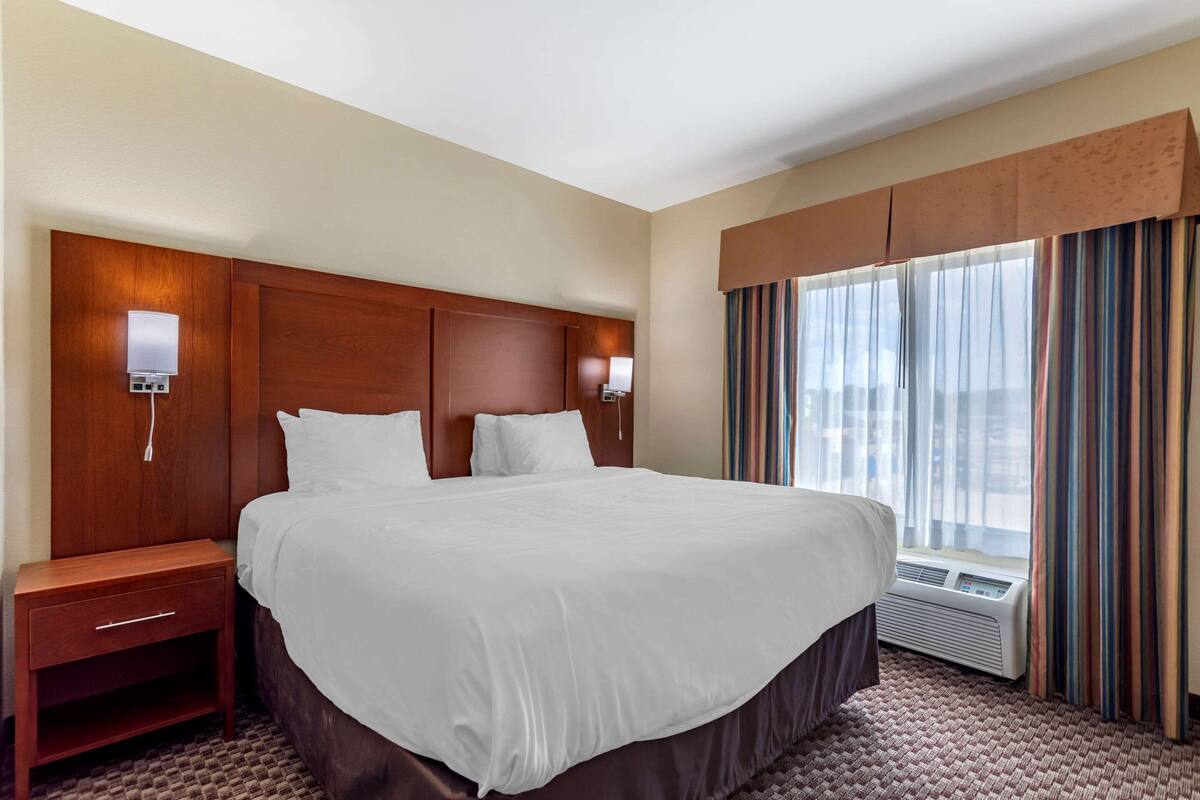A hotel room with a large bed, a nightstand, a wall-mounted lamp, and a window with curtains.