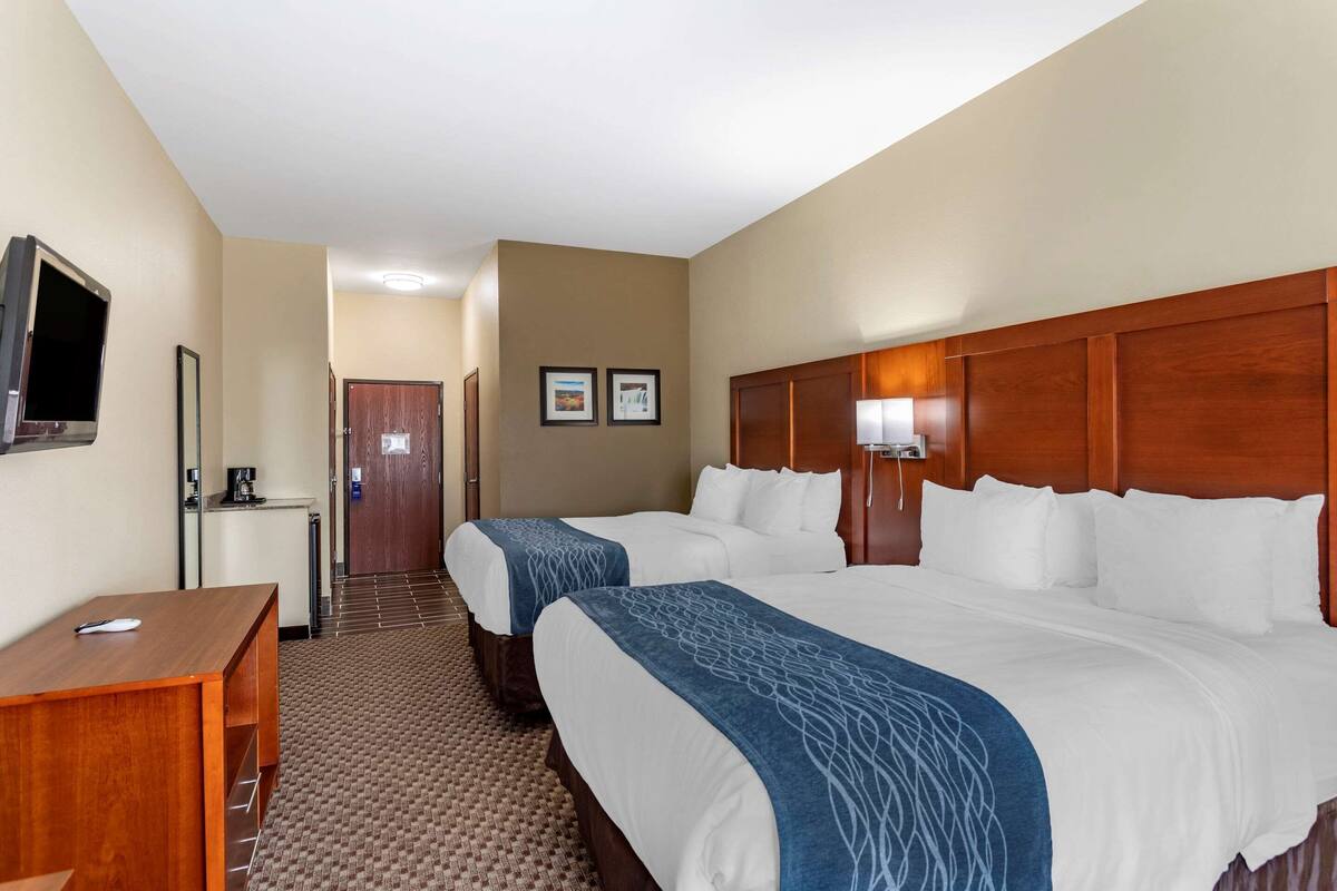A hotel room with two beds, a desk, a TV, and a door leading to a hallway.