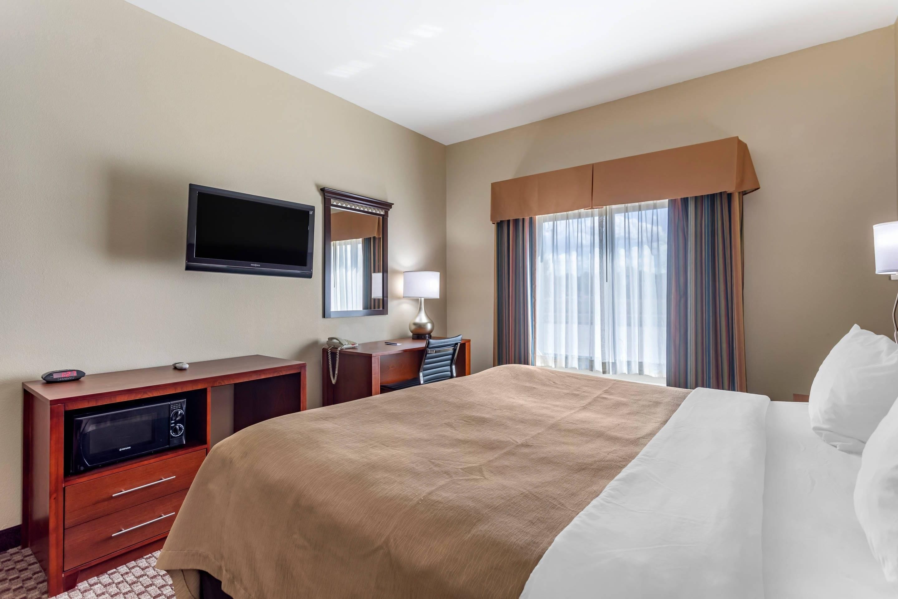 A hotel room with a large bed, a desk with a telephone, a flat-screen TV, a mirror, and a window with curtains.