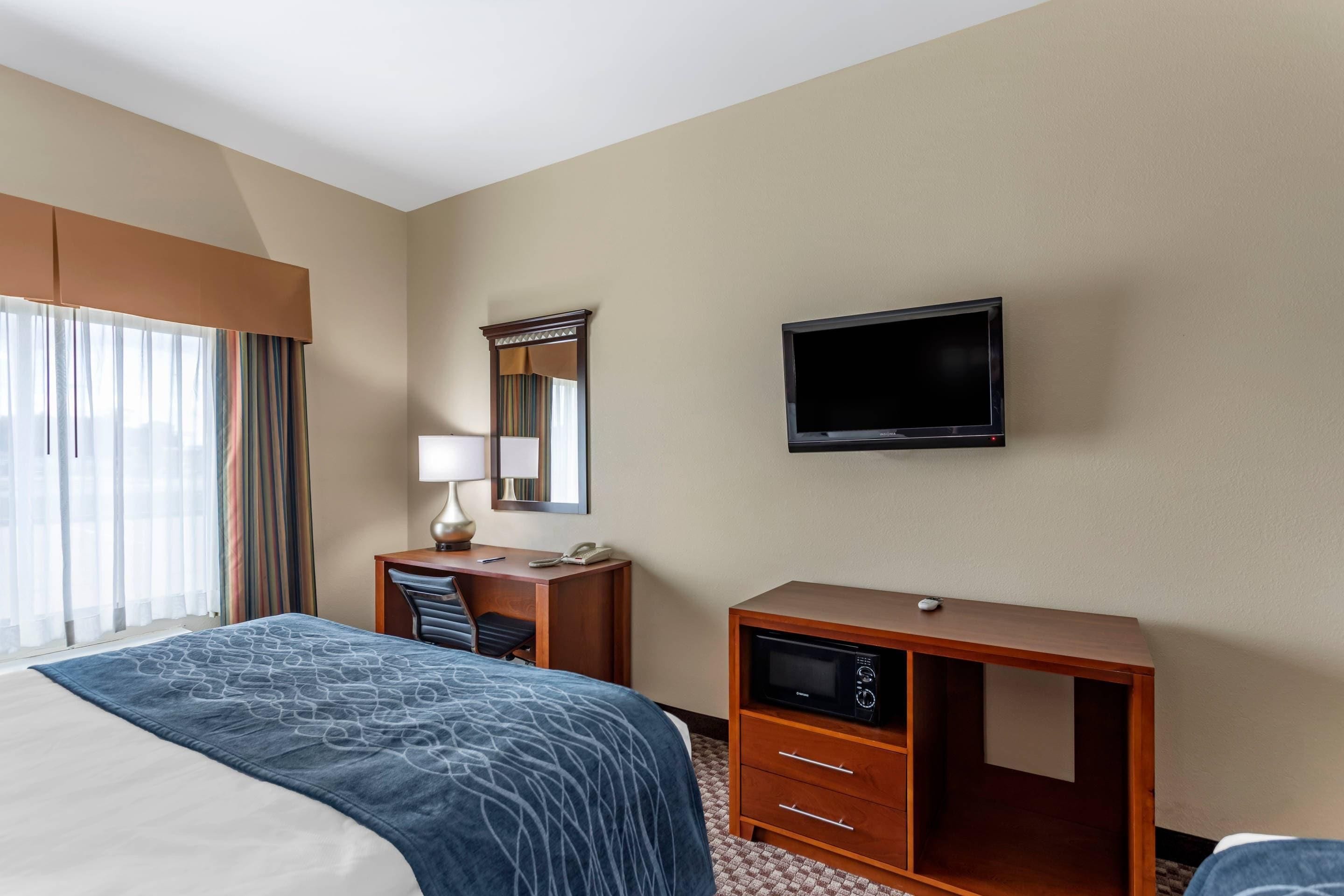 Quarto de hotel com cama, escrivaninha, cadeira, televisão e espelho.
