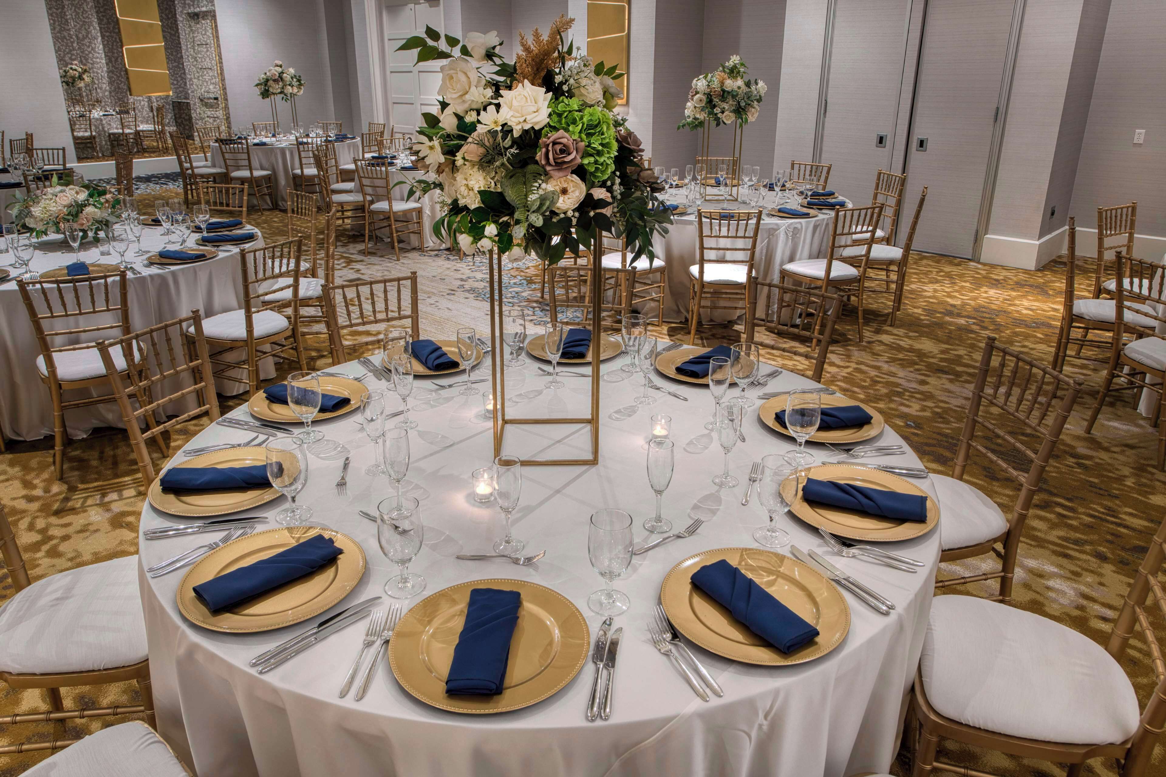 En festlokal med runda bord uppdukade för ett formellt evenemang, med guld- och vitfärgade dukar, blå servetter och blomsterarrangemang som mittpunkter.