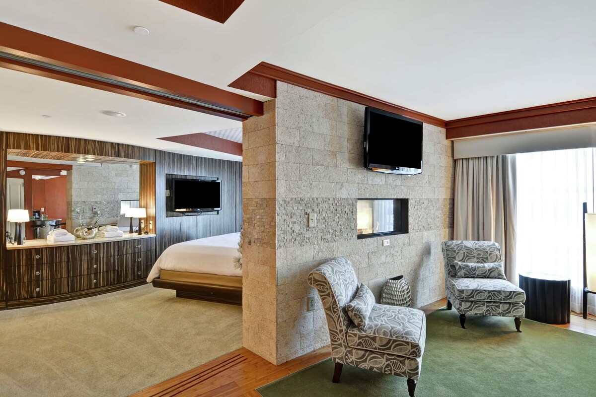 A hotel room with a bed, two patterned armchairs, a flat-screen TV mounted on a stone wall, a large mirror, and a window with curtains.