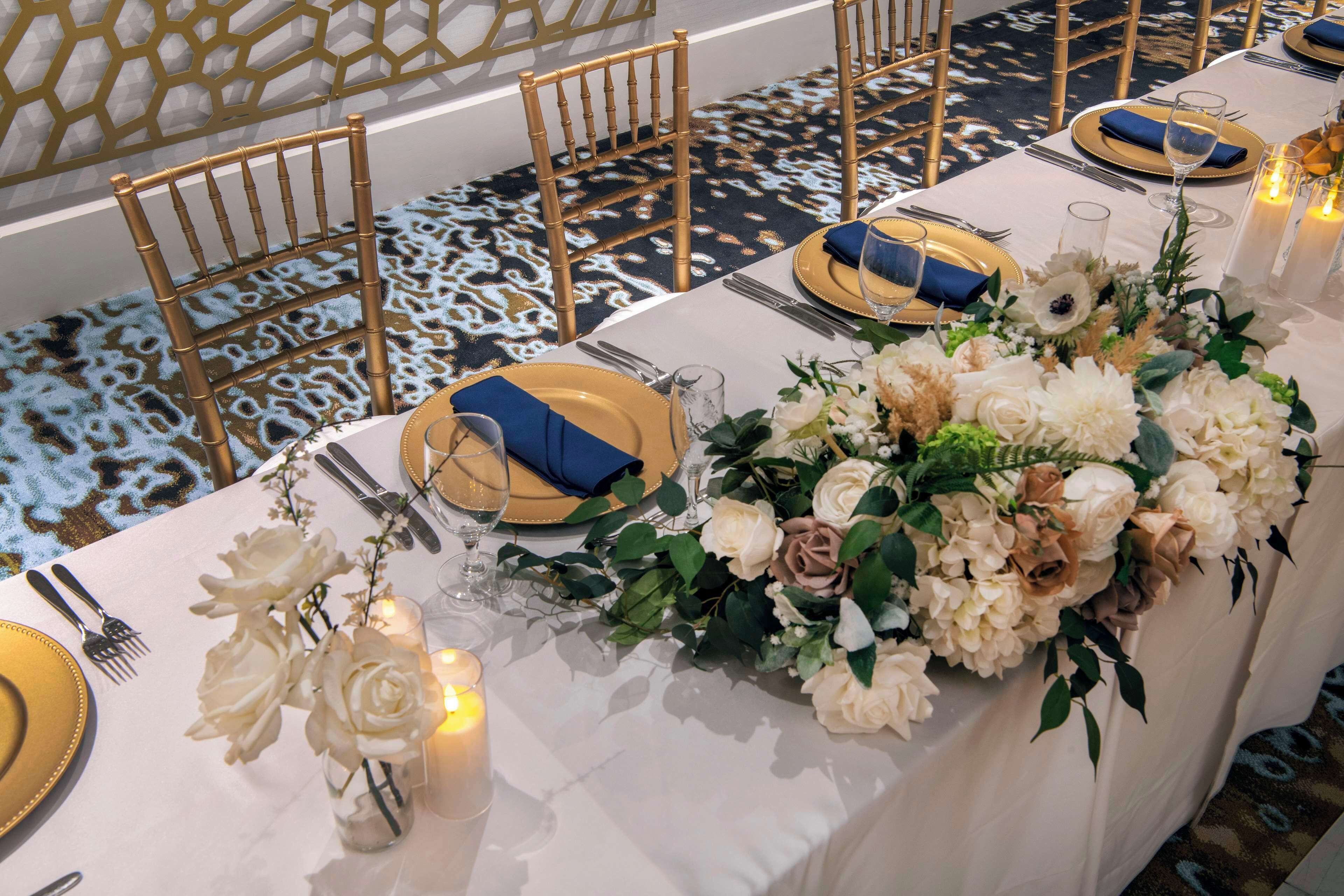 Ein Bankettisch mit gold- und blauem Geschirr, einer weißen Tischdecke und floralen Mittelpunkten.
