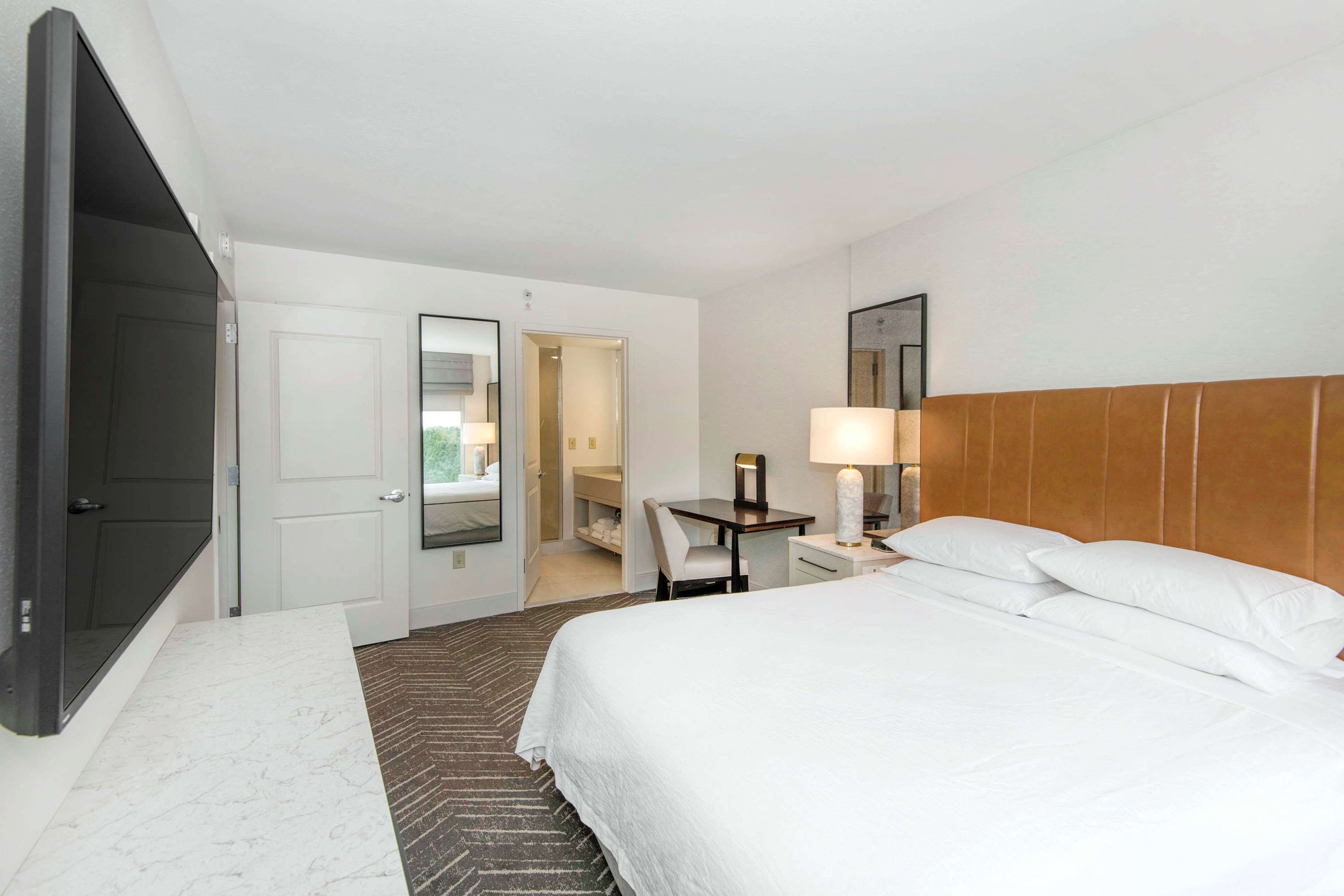 Una habitación de hotel con una cama, un escritorio, una silla, una lámpara, un espejo y una puerta.