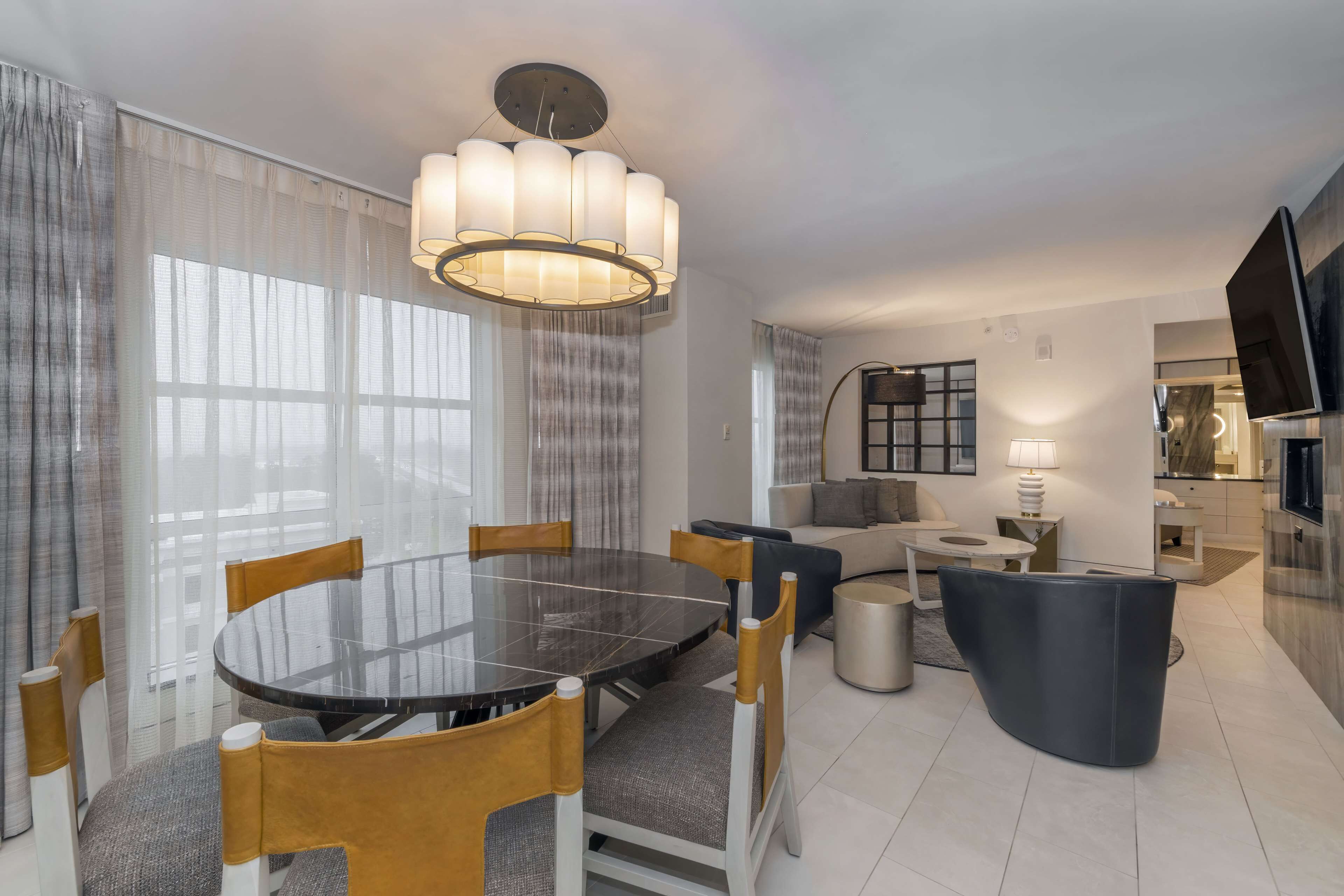 A modern living room with a round glass table, comfortable seating, a large chandelier, and a flat-screen TV.
