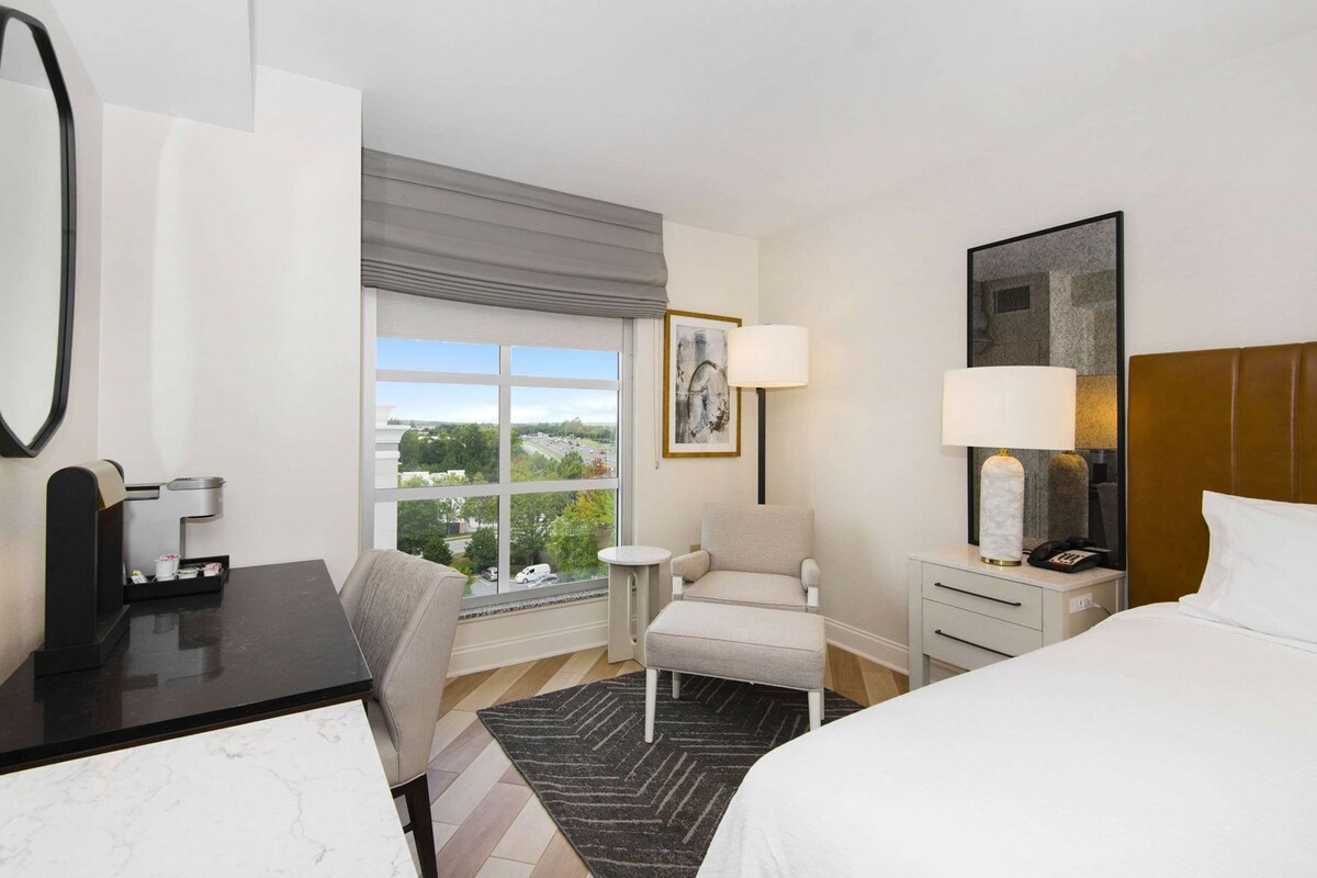 A hotel room with a bed, a desk, a chair, a nightstand, a lamp, a painting, and a window with a view of greenery and a road.