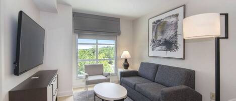 A modern living room with a flat-screen TV, grey sofa, round coffee table, and a large window with a view of greenery.