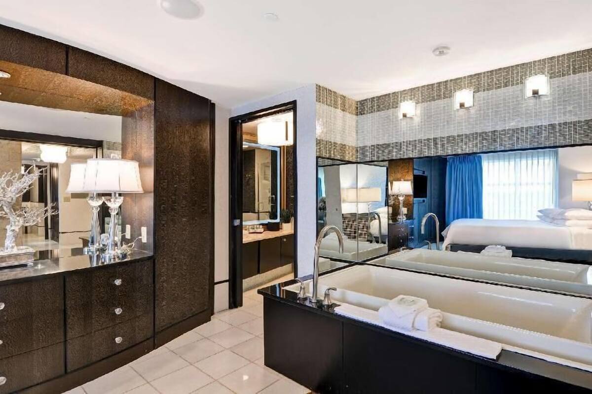 A modern hotel room with a large mirror, a bathtub, a vanity with a sink, and a bed.