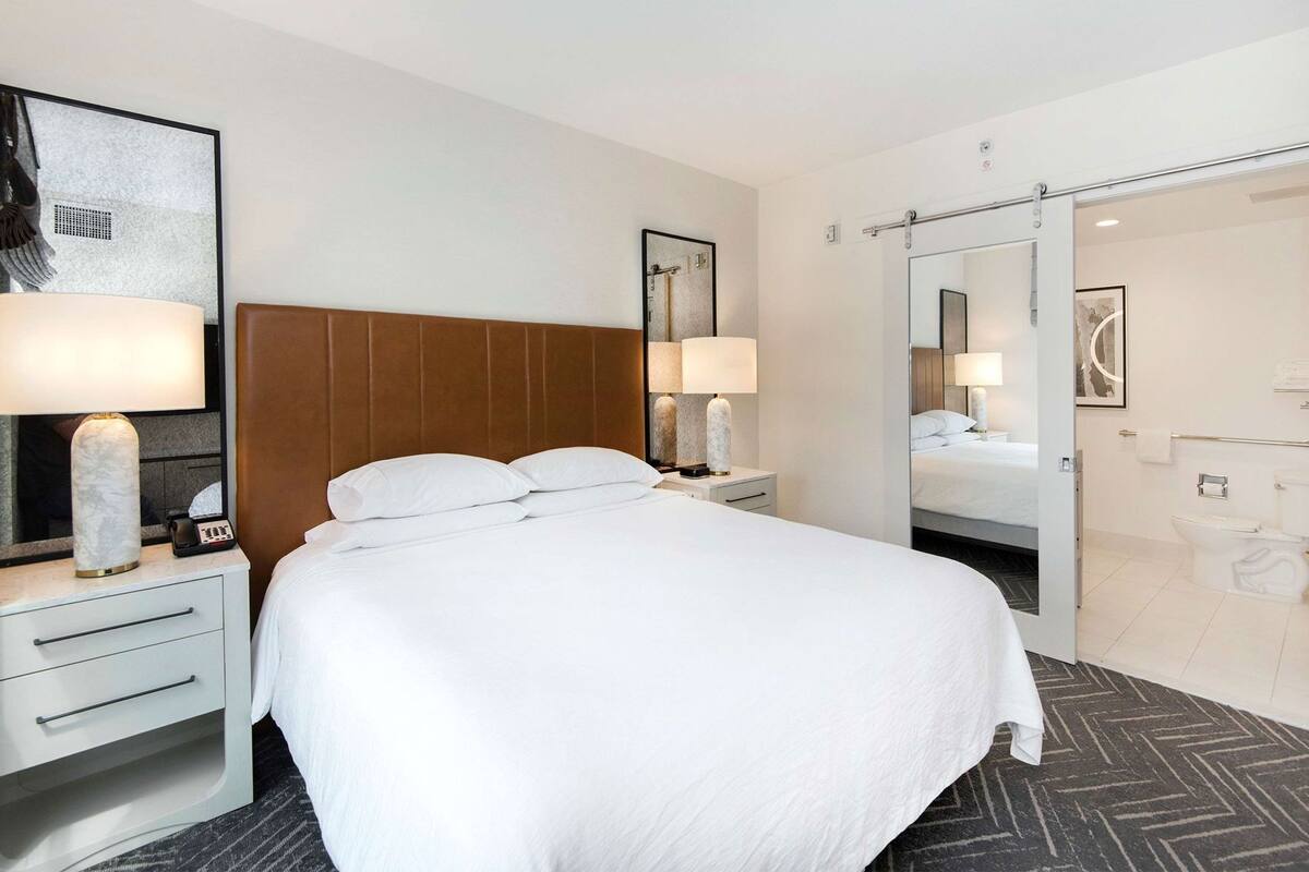 A hotel room with a large bed, a nightstand with a lamp, a mirror, and a bathroom with a toilet and sink.
