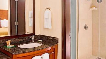 A hotel bathroom with a granite countertop sink, a mirror, a towel rack, and a shower area.