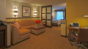 A hotel room with a sofa, a small table with a vase of flowers, a desk with a computer, and a bed in the background.