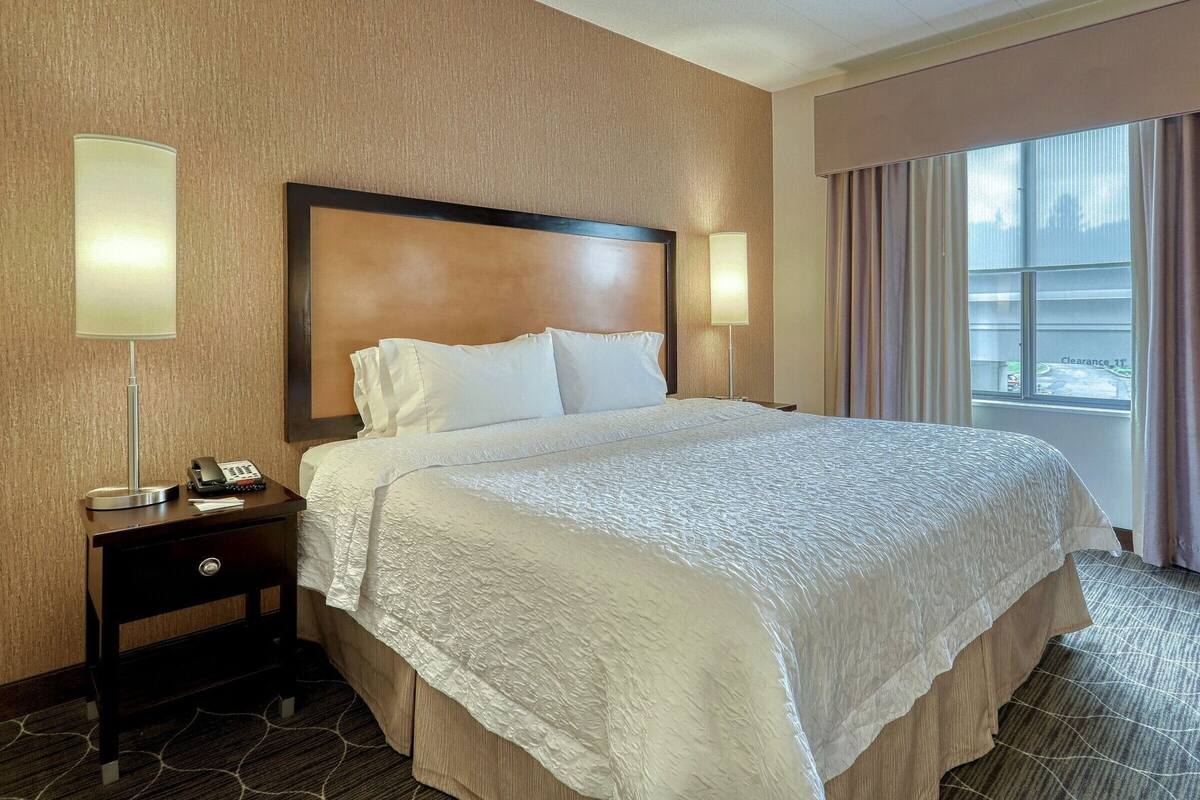 A hotel room with a large bed, a bedside table with a lamp, a phone, and a window with curtains.