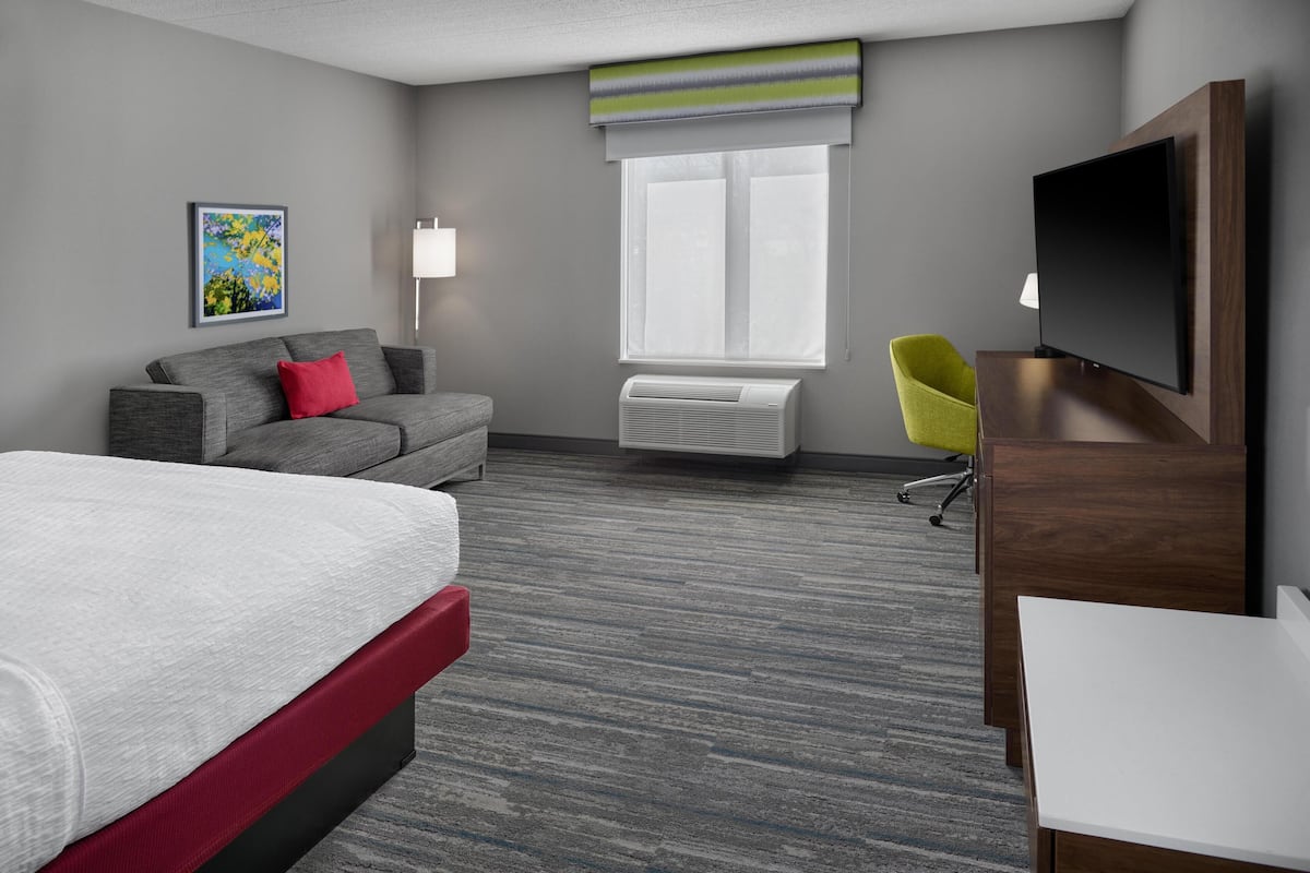 A hotel room with a bed, a grey sofa, a yellow chair, a TV, and a window with blinds.