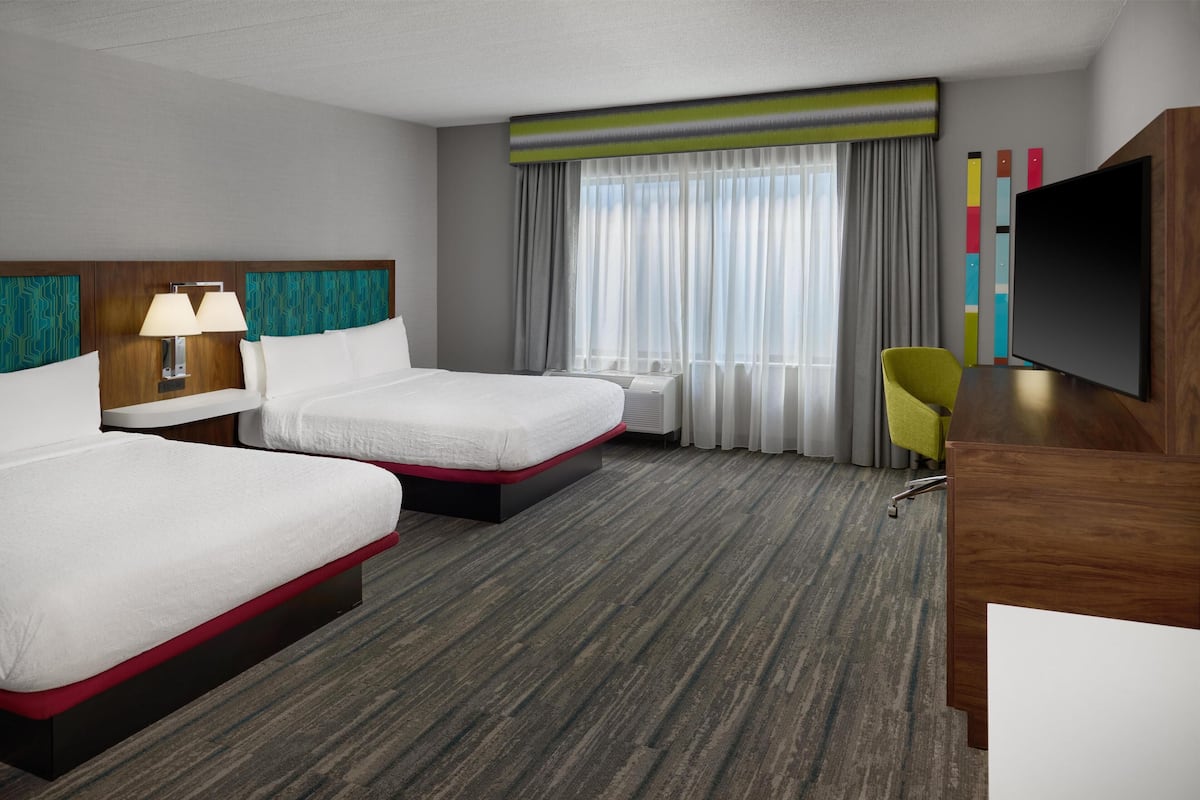 A hotel room with two beds, a desk, a chair, a TV, and a large window with curtains.