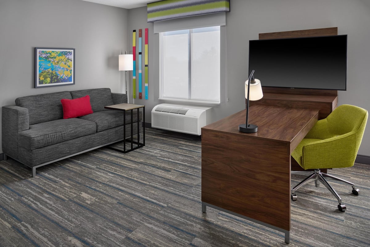 A modern hotel room with a grey sofa, a red pillow, a wooden desk, a green chair, a flat-screen TV, a standing lamp, a colorful abstract painting, a window with blinds, and a built-in air conditioner.