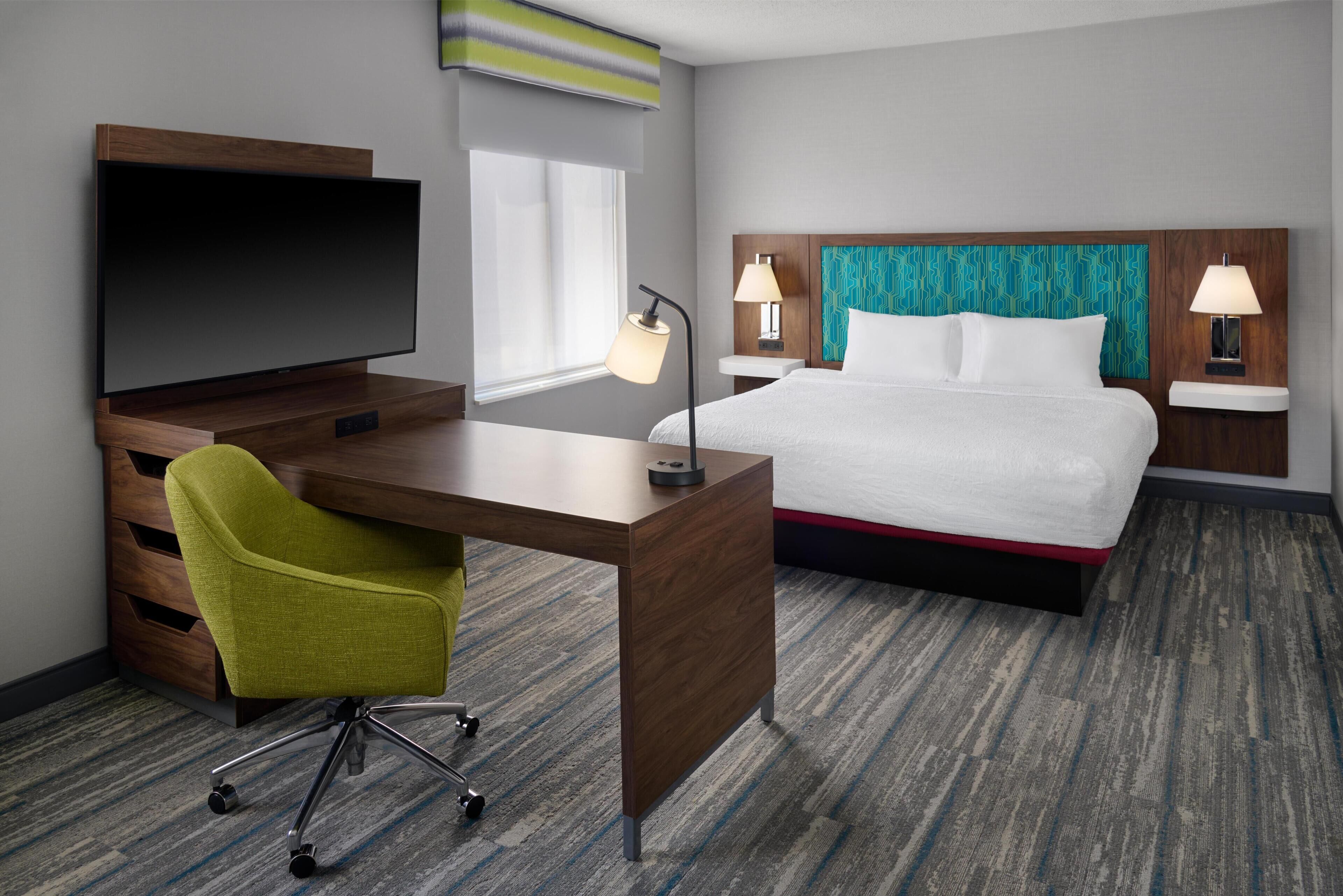 A hotel room with a bed, desk, chair, TV, lamps, and a patterned carpet.