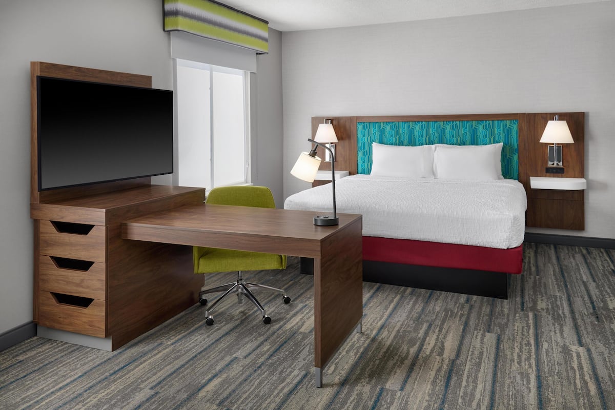 A hotel room with a wooden desk, a flat-screen TV, a bed with a teal headboard, two bedside lamps, and a green chair.