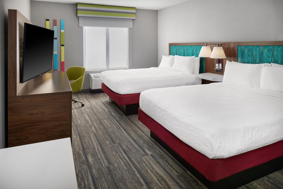 A hotel room with two beds, a TV, a desk, a chair, and a window with blinds.