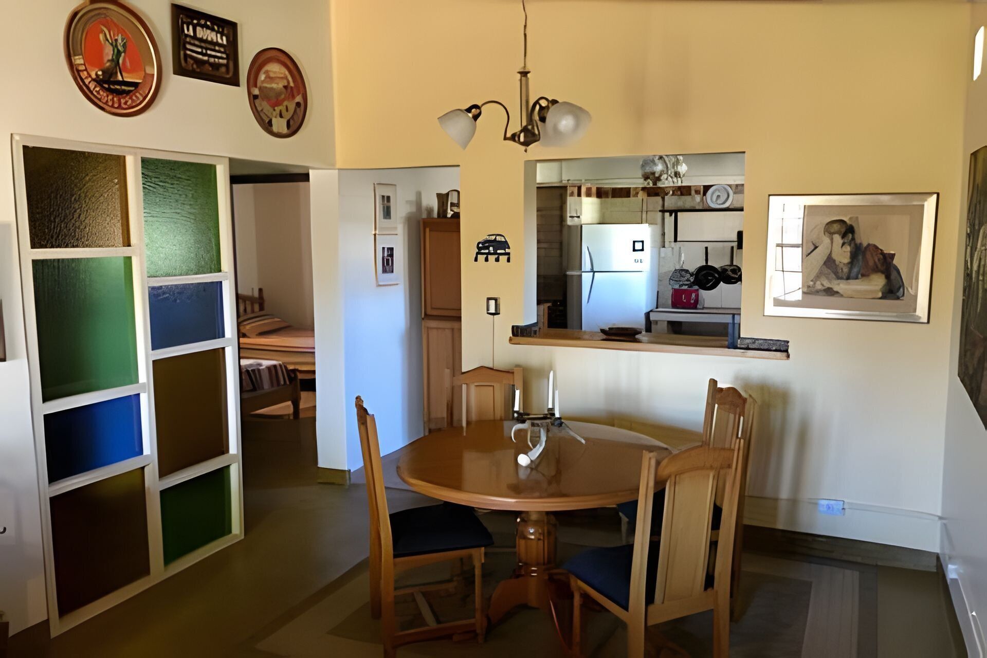Un coin repas avec une table ronde, quatre chaises et un lustre. On aperçoit une cuisine à l’arrière-plan, avec un réfrigérateur et une fenêtre.