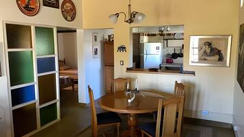 Un coin repas avec une table ronde, quatre chaises et un lustre. On aperçoit une cuisine à l’arrière-plan, avec un réfrigérateur et une fenêtre.