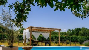 Area piscina con una pergola rivestita di tessuto bianco, una struttura in pietra e un cielo azzurro limpido.