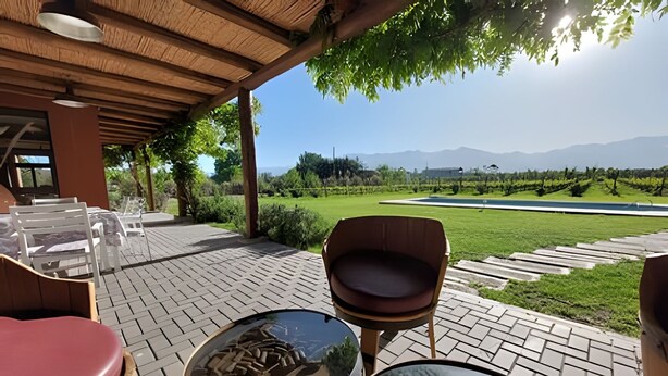 Area esterna con sedute, tavoli, una pergola in legno e vista su un giardino curato e montagne sullo sfondo.