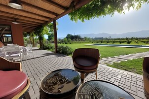 Uma área externa com assentos, cadeiras e mesas, uma pérgola de madeira e vista para um jardim bem cuidado e montanhas ao longe.