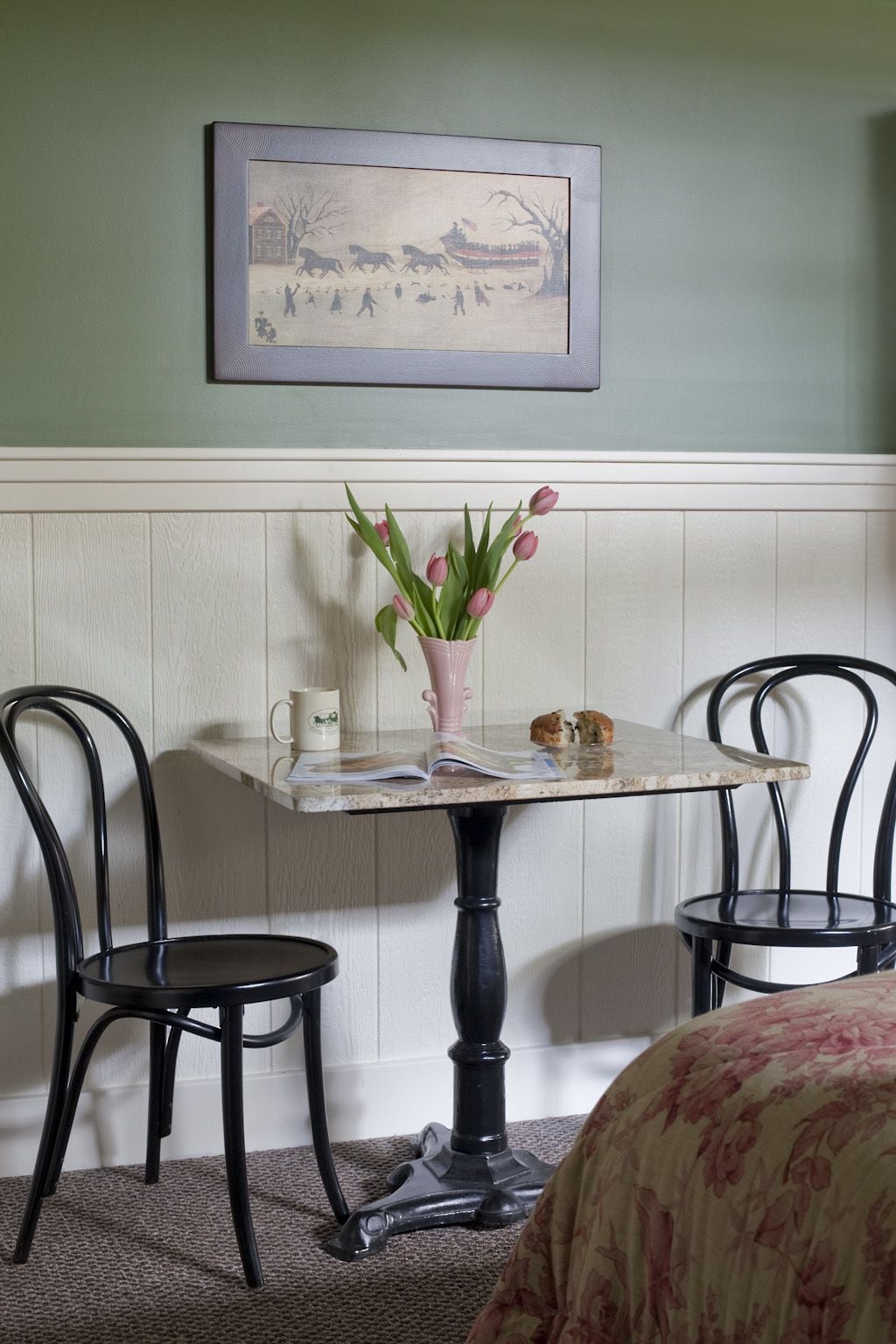 Un comedor con una mesa, sillas, un jarrón de tulipanes, un cuadro en la pared y una mesita con una taza y un pastel.