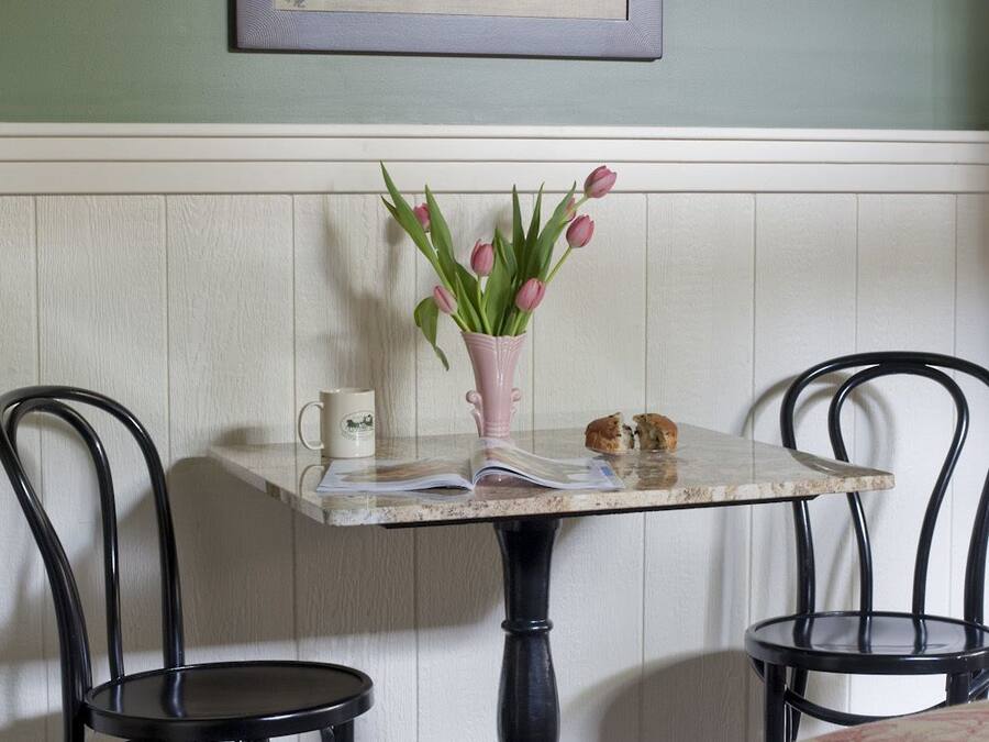 Un comedor con una mesa, sillas, un jarrón de tulipanes, un cuadro en la pared y una mesita con una taza y un pastel.