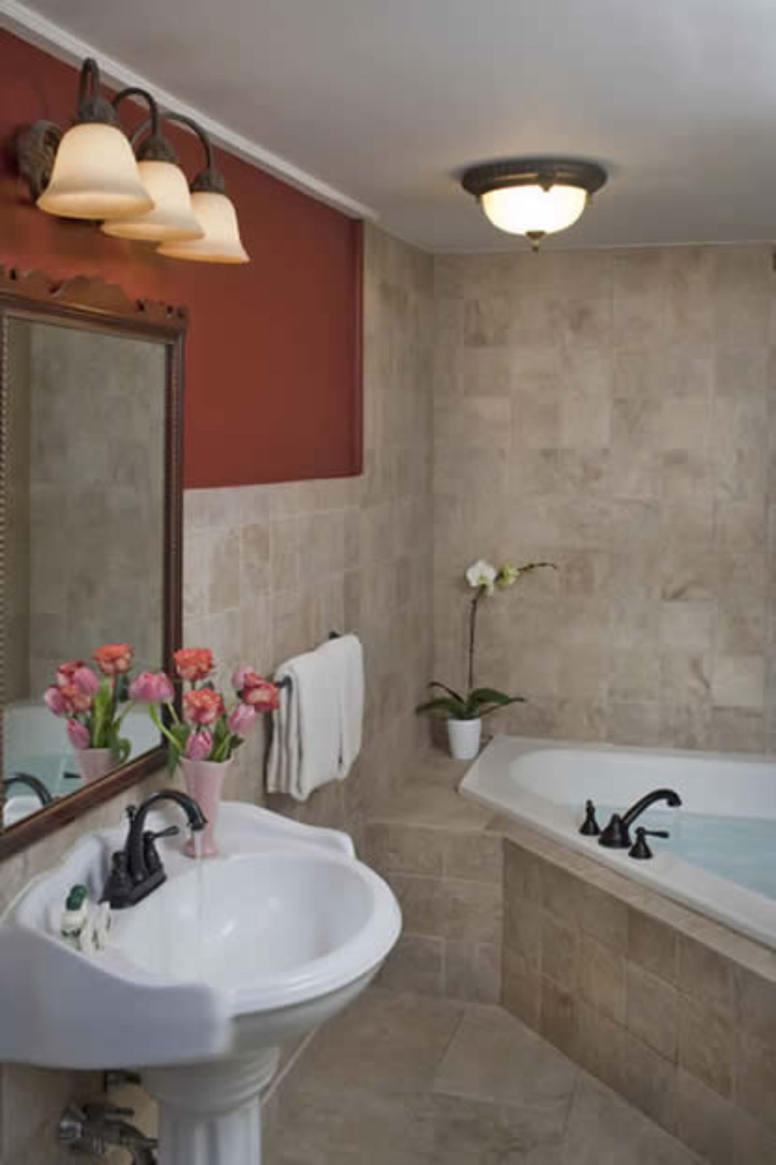 A bathroom with a white sink, a large mirror, a wall-mounted light fixture, a bathtub, and a potted orchid.