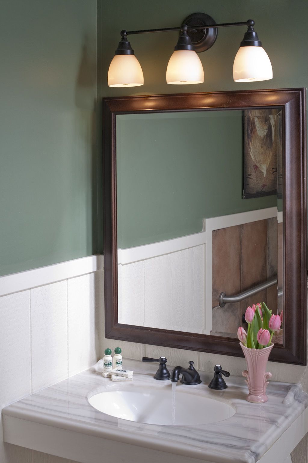 Un baño con un lavamanos blanco, tres luces empotradas en la pared, un espejo grande y un florero con flores rosas.