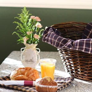 Mesa com um croissant, muffin e sumo de laranja, uma cesta de vime e um vaso com flores.