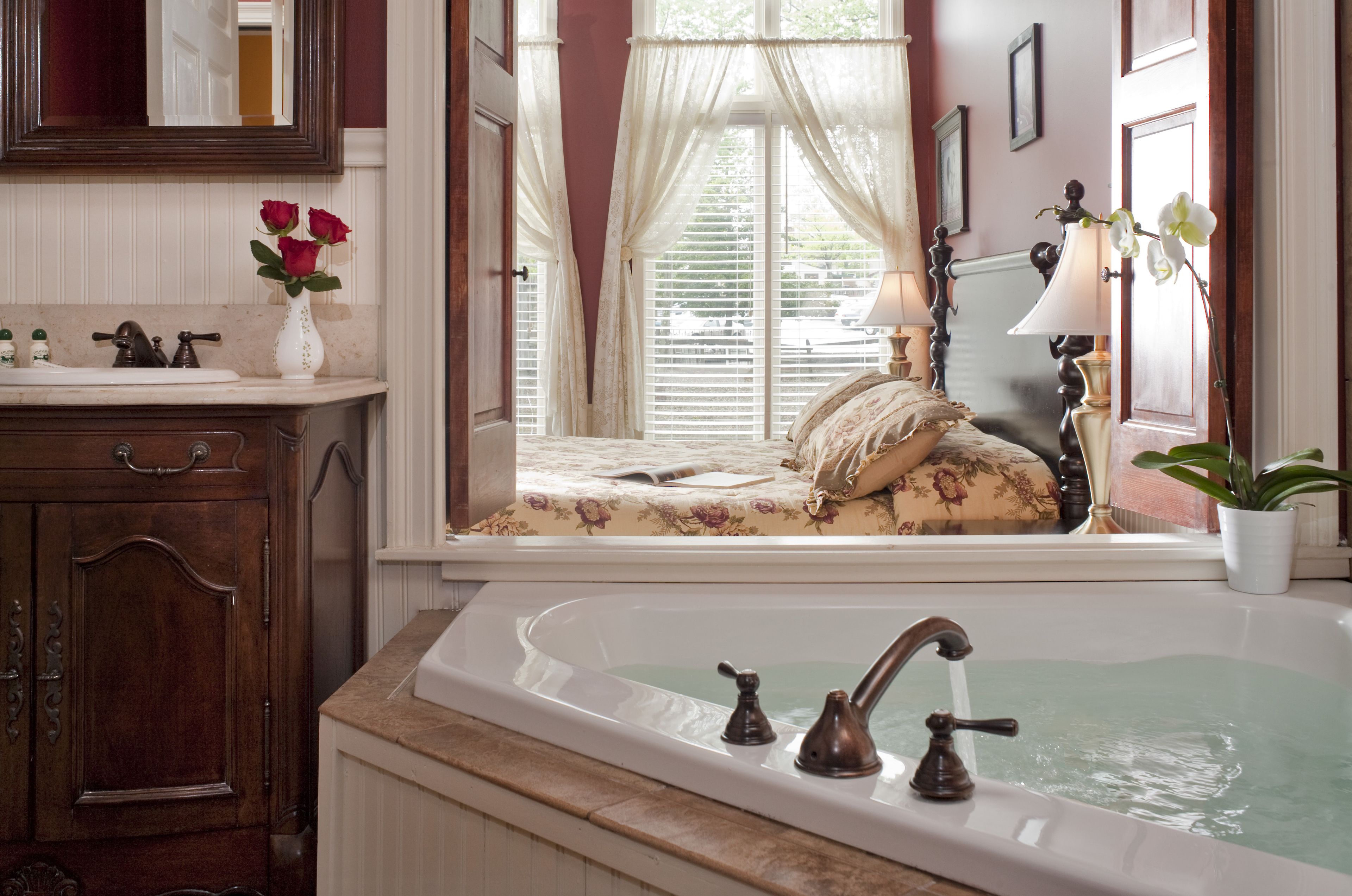 A bathroom with a bathtub, a vanity with a mirror, a window with curtains, and a bedside table with a lamp.