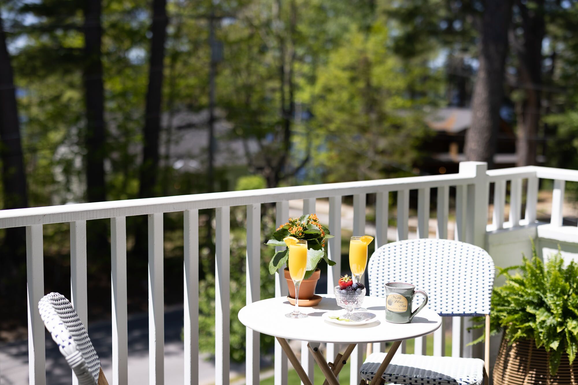 A small round table with a white tablecloth, two glasses of orange juice, a bowl of fruit, and a mug on a balcony with white railings.