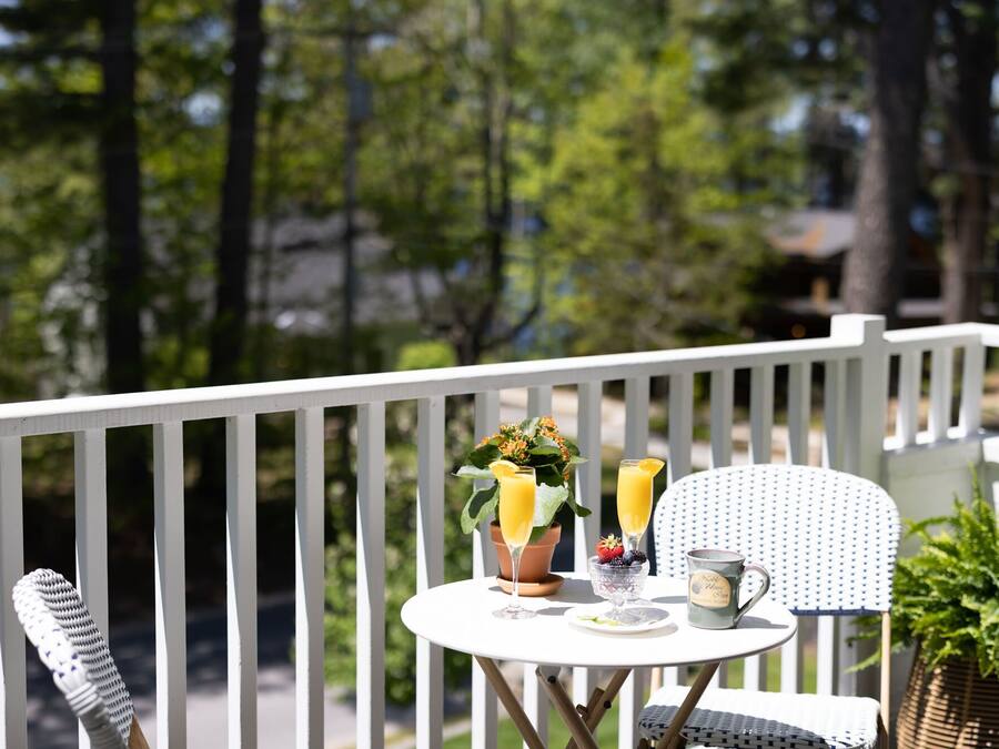 A small round table with a white tablecloth, two glasses of orange juice, a bowl of fruit, and a mug on a balcony with white railings.