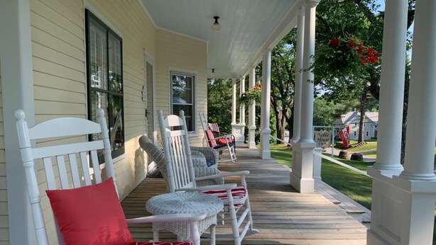 A porch with white wicker chairs and a red cushion, a wooden deck, and a sign for the Noble House Inn in the background. - Noble House Inn (Bridgton)