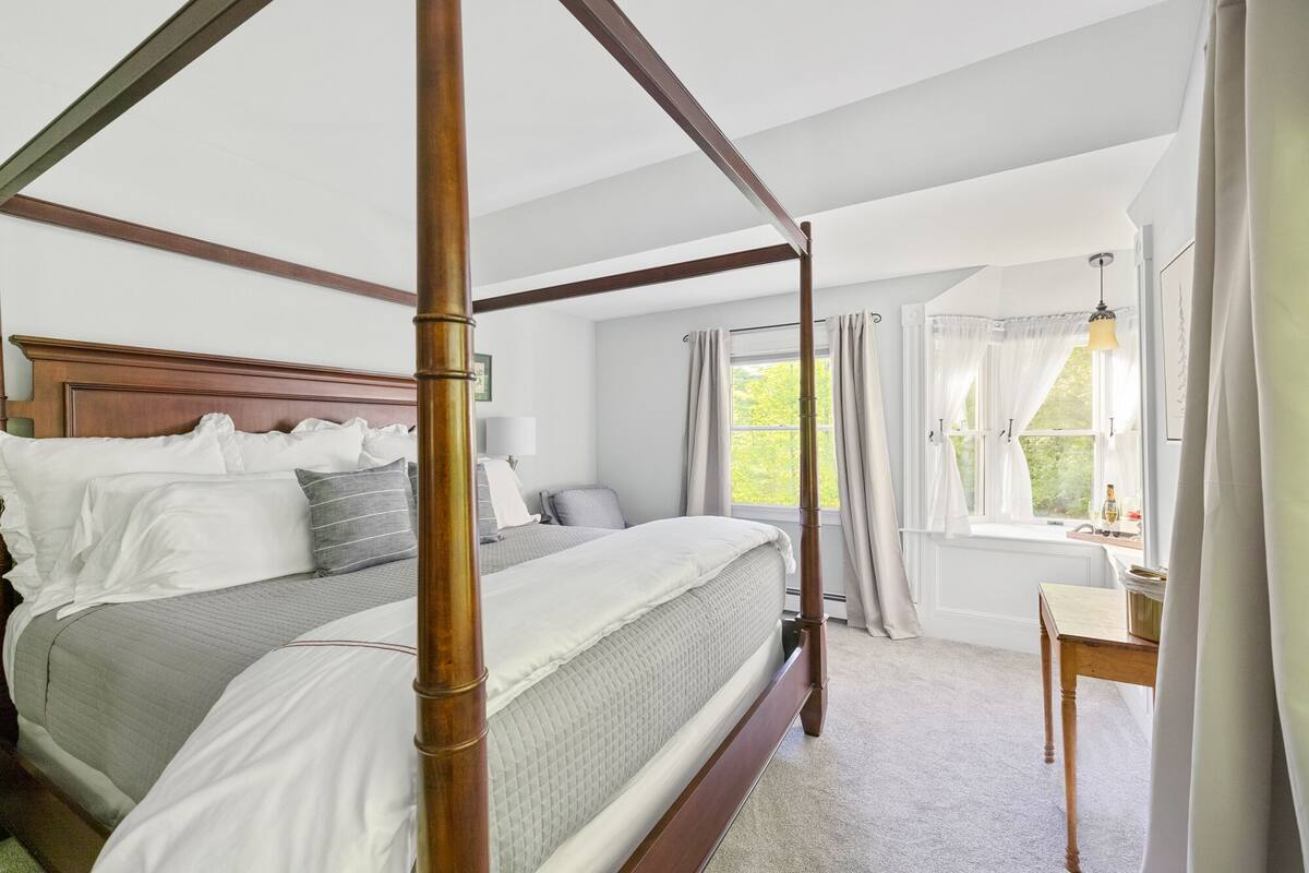 A four-poster bed with a wooden headboard, a bedside table with a lamp, and a window with curtains.