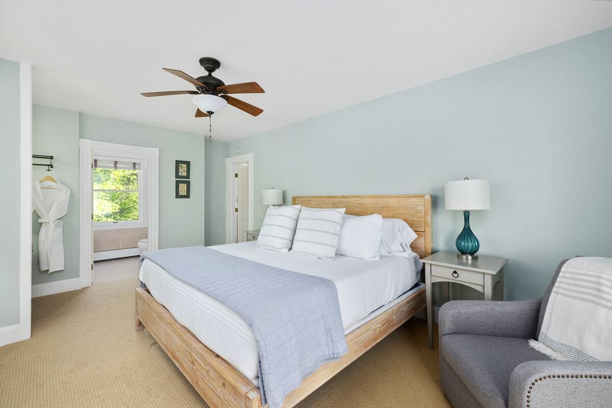A bedroom with a wooden bed, a ceiling fan, a nightstand with a lamp, a chair, and a view of the outdoors.