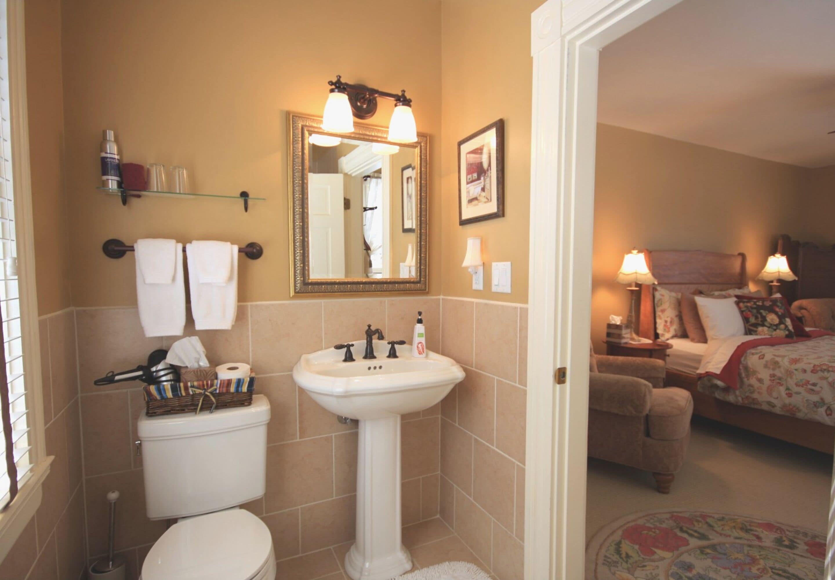 A bathroom with a toilet, sink, and mirror. There is a bed in the adjacent room.