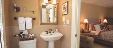 A bathroom with a toilet, sink, and mirror. There is a bed in the adjacent room.