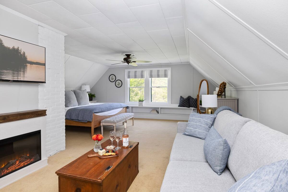 A cozy attic bedroom with a bed, a sitting area with a sofa, a fireplace, and a window with curtains.
