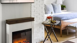A bedroom with a fireplace, a bed, a bedside table with a bottle and flowers, and a view of a lake. - Noble House Inn (Bridgton)