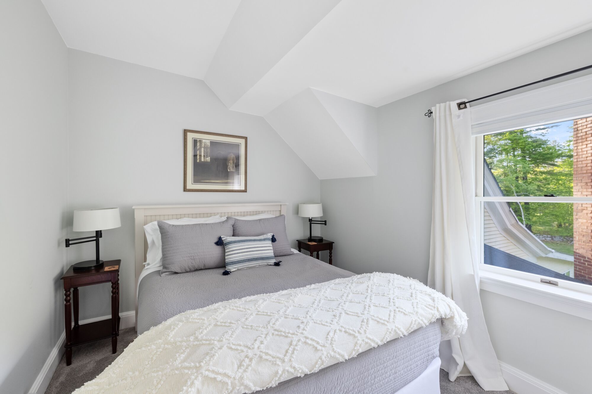 A bedroom with a bed, two bedside tables, a nightstand, a framed picture, and a window with curtains.