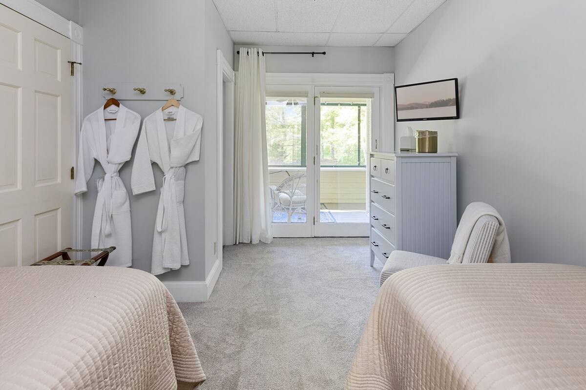 A bedroom with two beds, a white wardrobe, a desk, and a window with a view of greenery.
